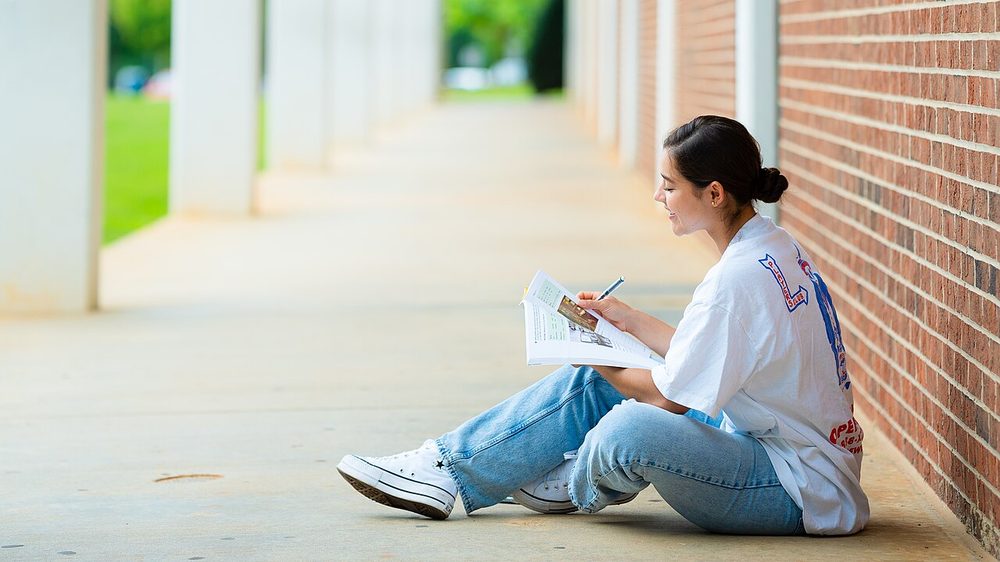 A student sitting on the floor of a corridor reading a workbook